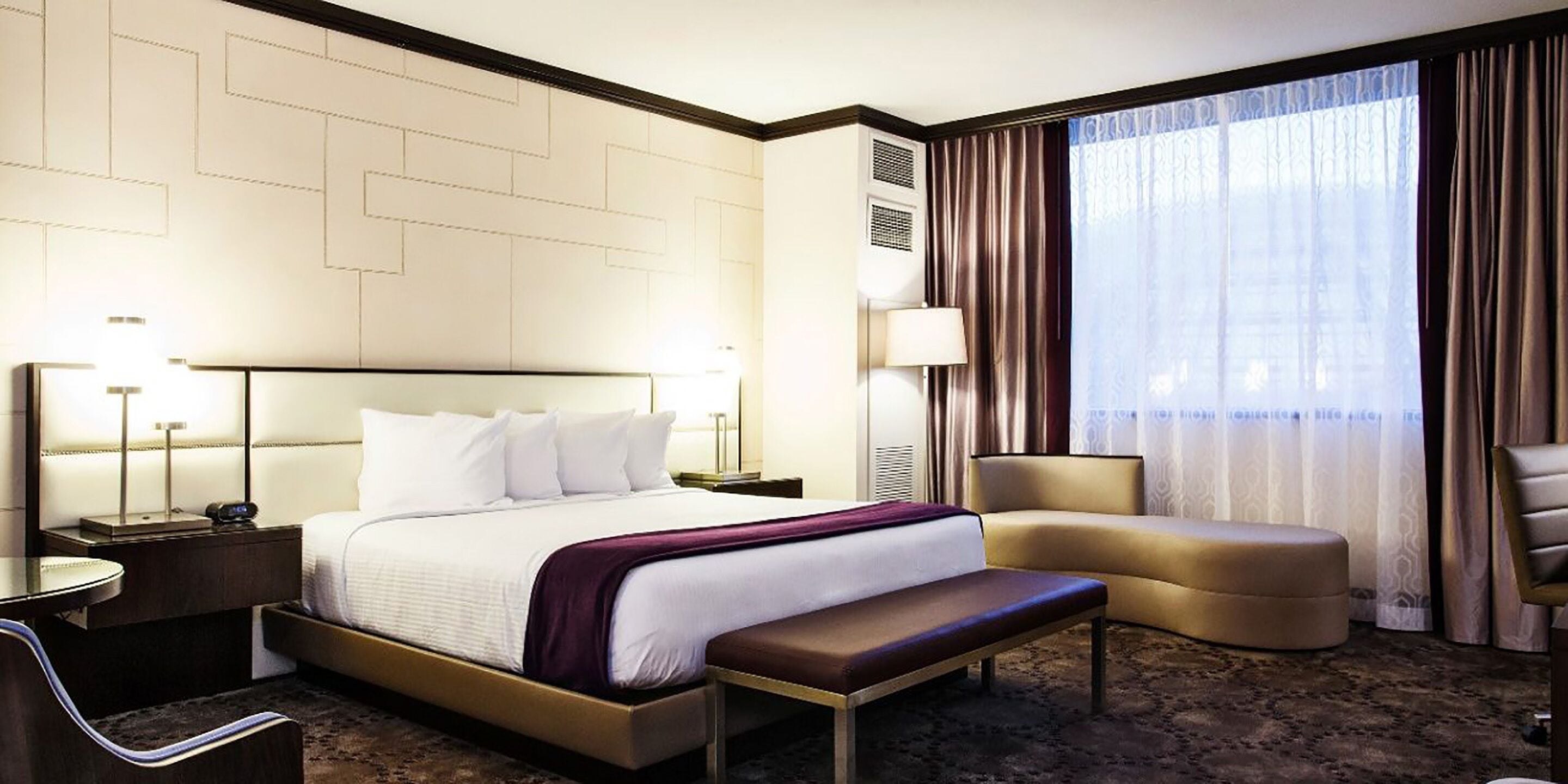 A hotel room with a large bed, a bedside table with a lamp, a chair, a bench, and a large window with curtains.