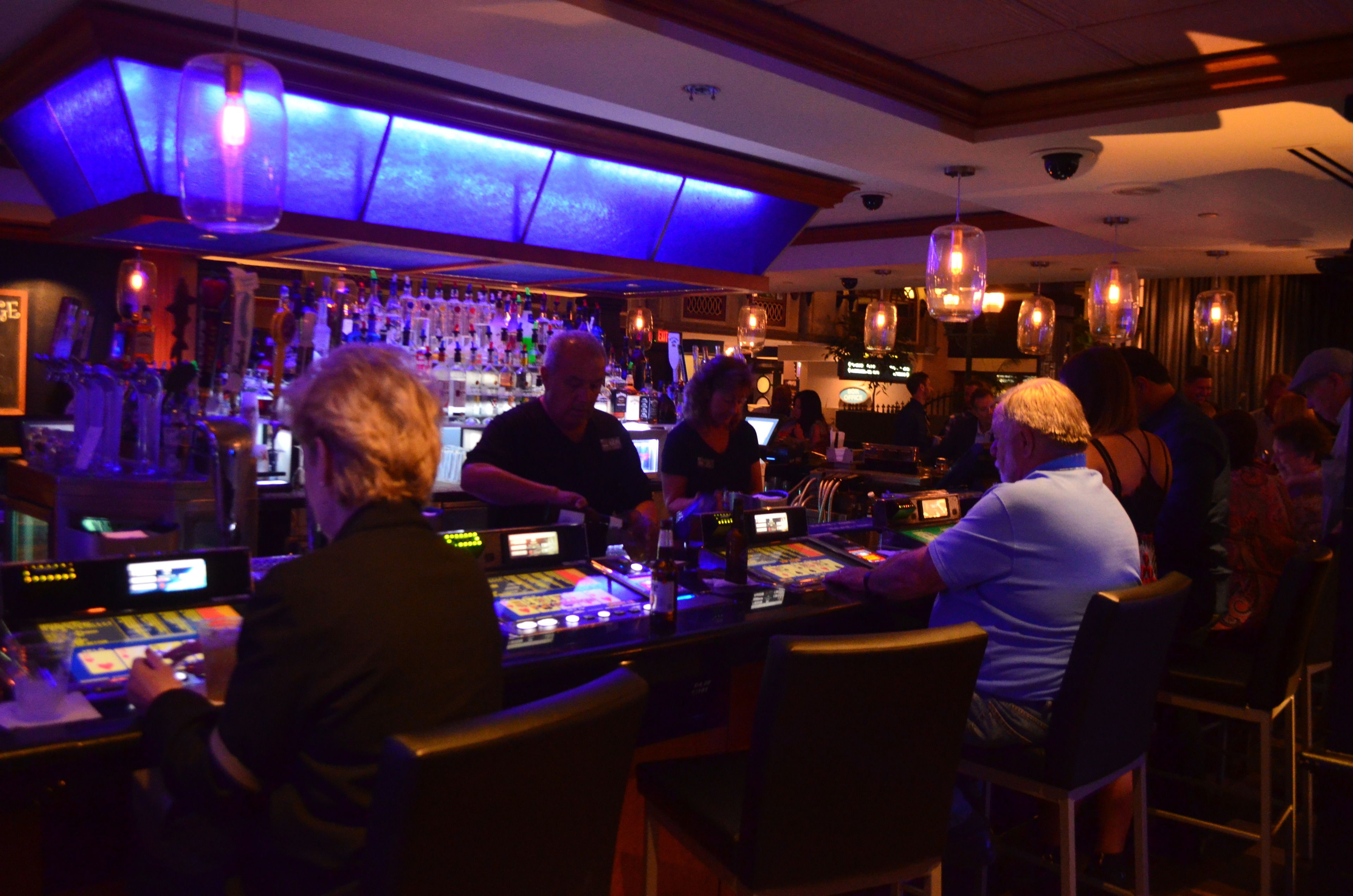 Zona de bar con varios clientes sentados en mesas, barra con distintas botellas e iluminación colgante.