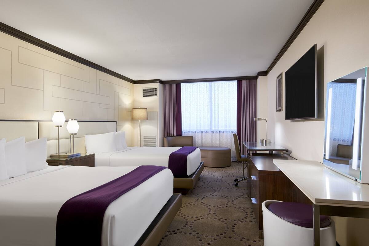 Ein Hotelzimmer mit zwei Betten, einem Schreibtisch, einem Stuhl, einem Fernseher und einem Fenster mit Vorhängen.