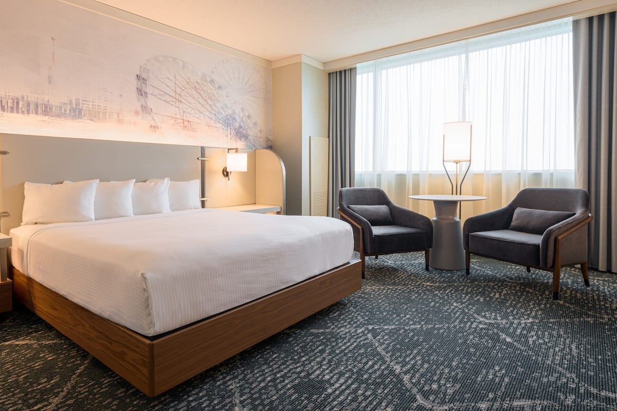 Ein Hotelzimmer mit einem großen Bett, zwei Sesseln, einem kleinen Tisch und einem großen Fenster mit Vorhängen.