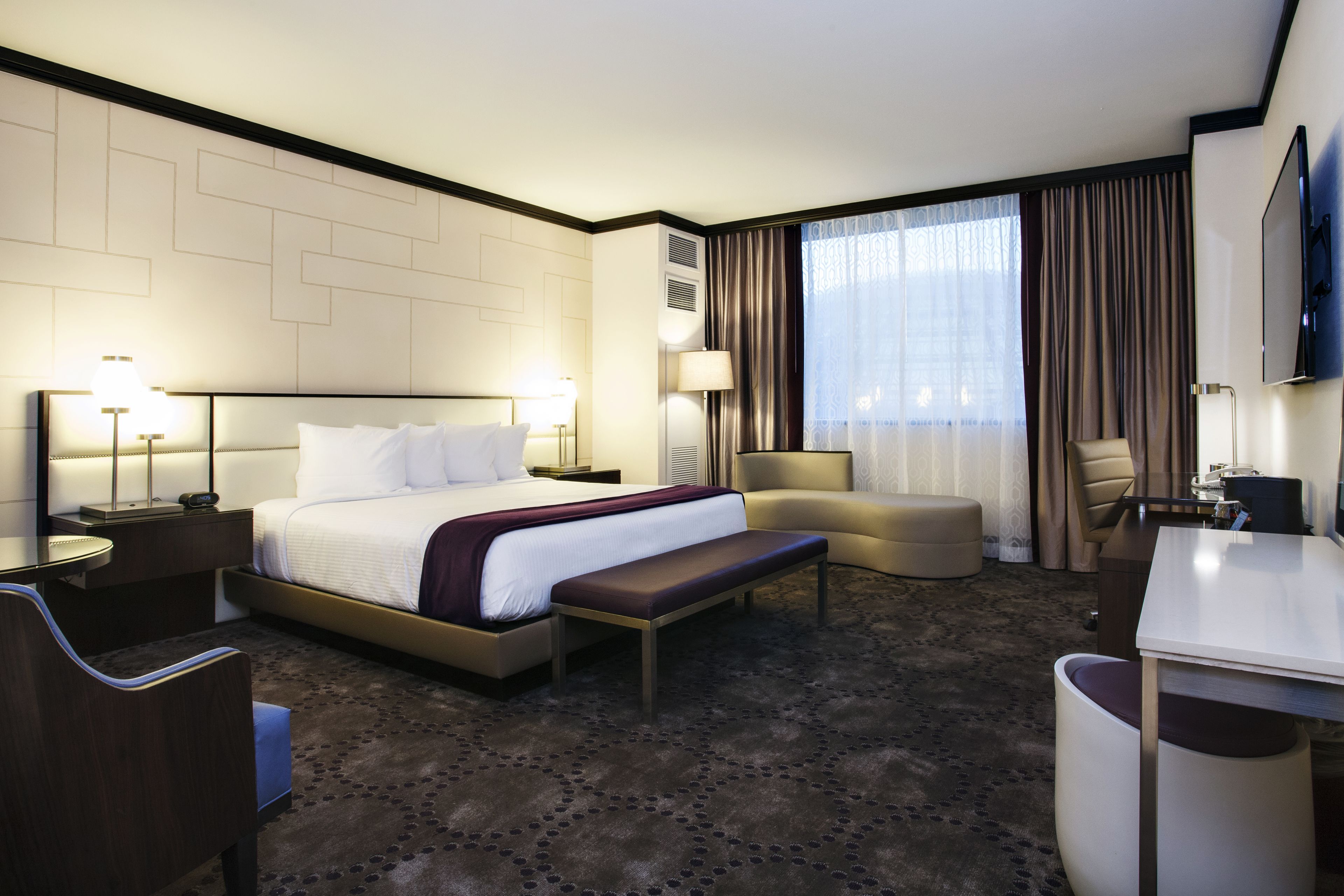 A hotel room with a large bed, a desk, a chair, a bench, a TV, and a window with curtains.