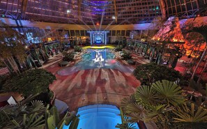 Amplia zona de piscina interior con un escenario central, rodeada de asientos y exuberante vegetación.