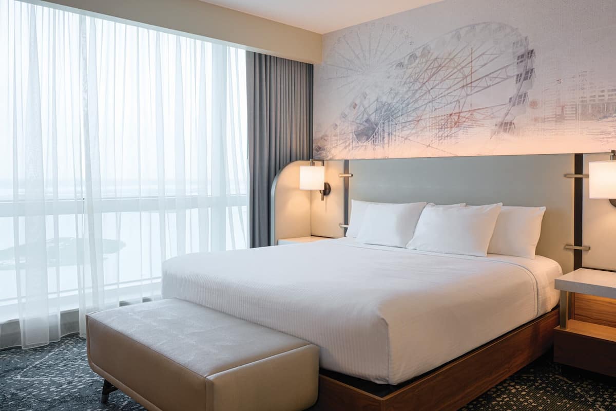 Ein Hotelzimmer mit einem großen Bett, einer Bank, zwei Nachttischen, einer Wandlampe und einem großen Fenster mit Vorhängen.