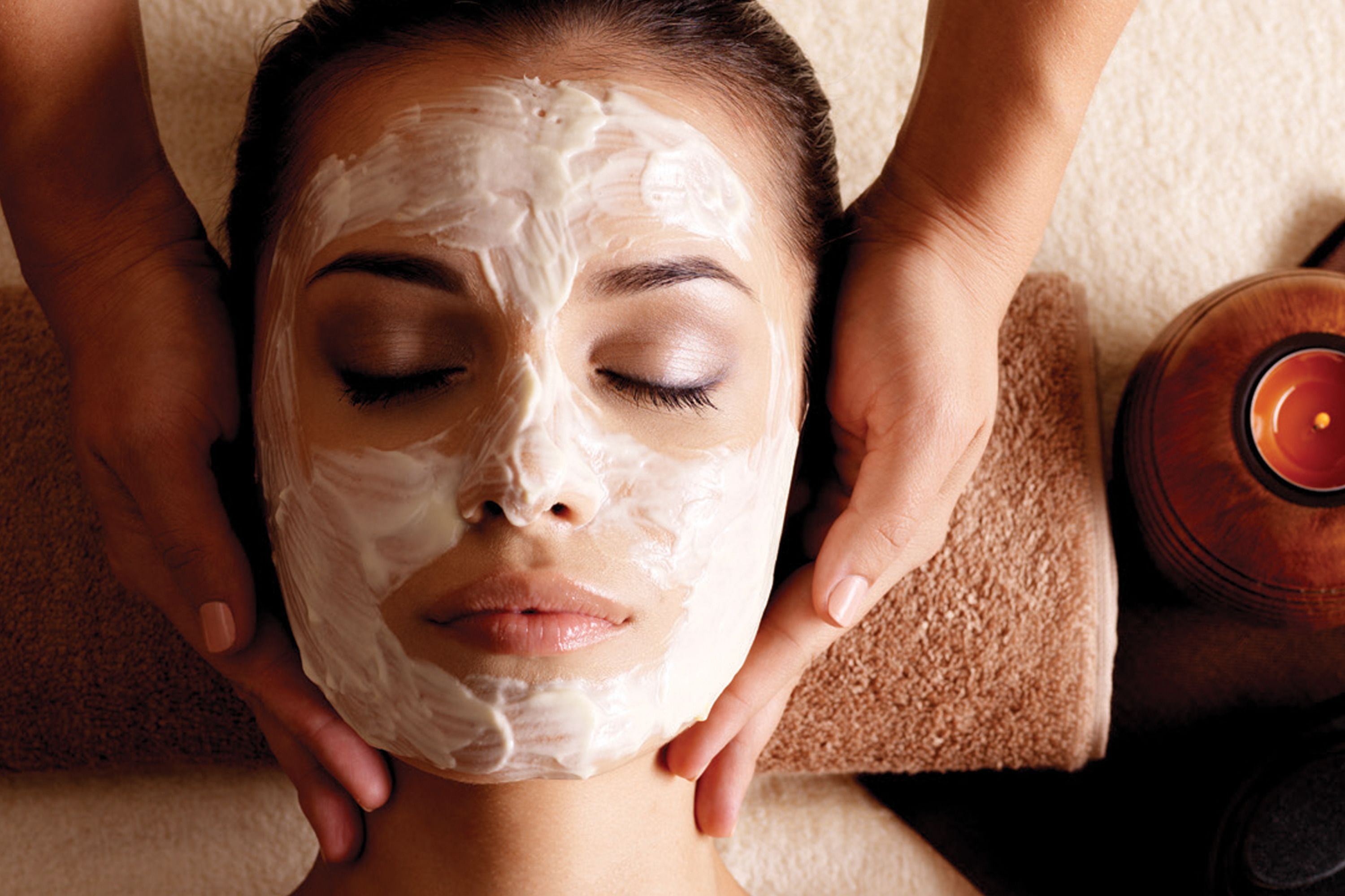 Una persona recibiendo un tratamiento facial con una toalla y una vela sobre una mesa.