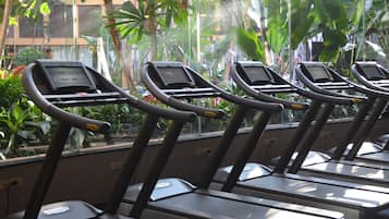 A row of treadmills with digital displays, positioned near a window with a view of lush greenery and a building in the background.