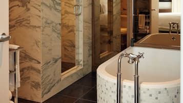A modern bathroom with a marble wall, a glass-enclosed shower, a freestanding bathtub, and a mirror reflecting the room.