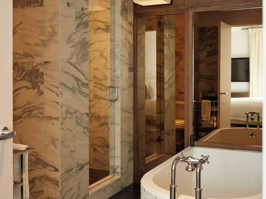 A modern bathroom with a marble wall, a glass-enclosed shower, a freestanding bathtub, and a mirror reflecting the room.