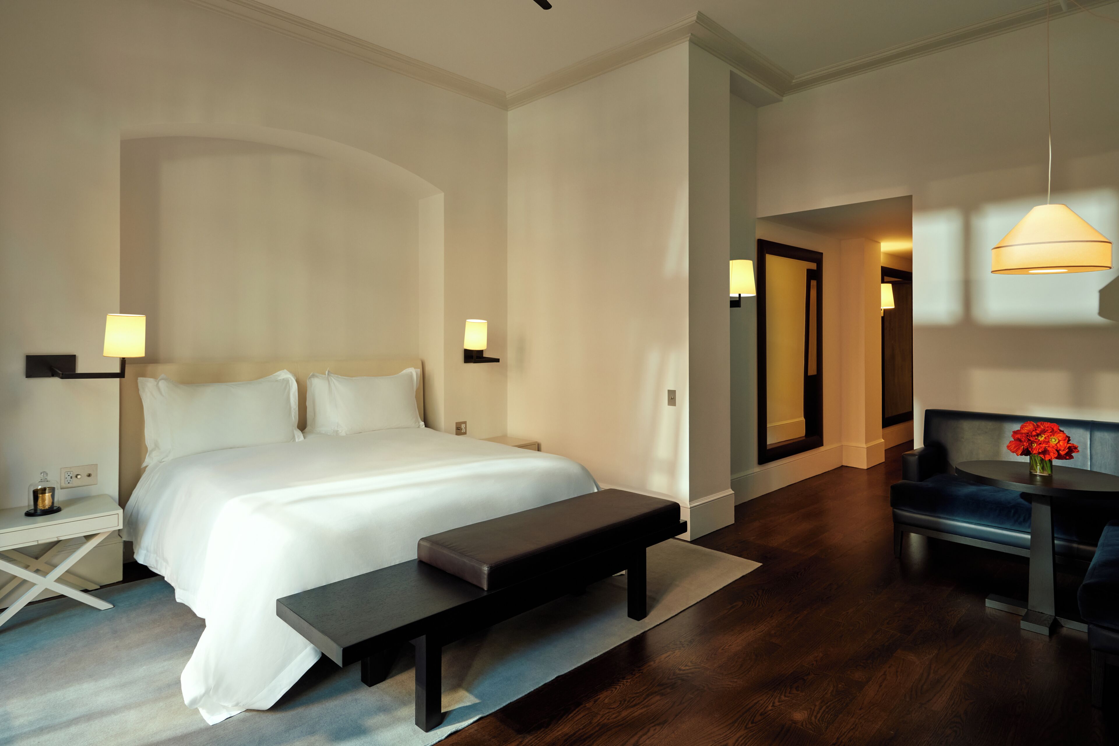 A hotel room with a large bed, bedside tables, a bench, a desk, and a seating area with a lamp.