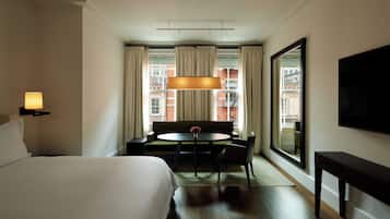 Une chambre d’hôtel comprenant un lit, une table à manger avec des chaises, un canapé, une grande fenêtre avec des rideaux et un téléviseur à écran plat fixé au mur.