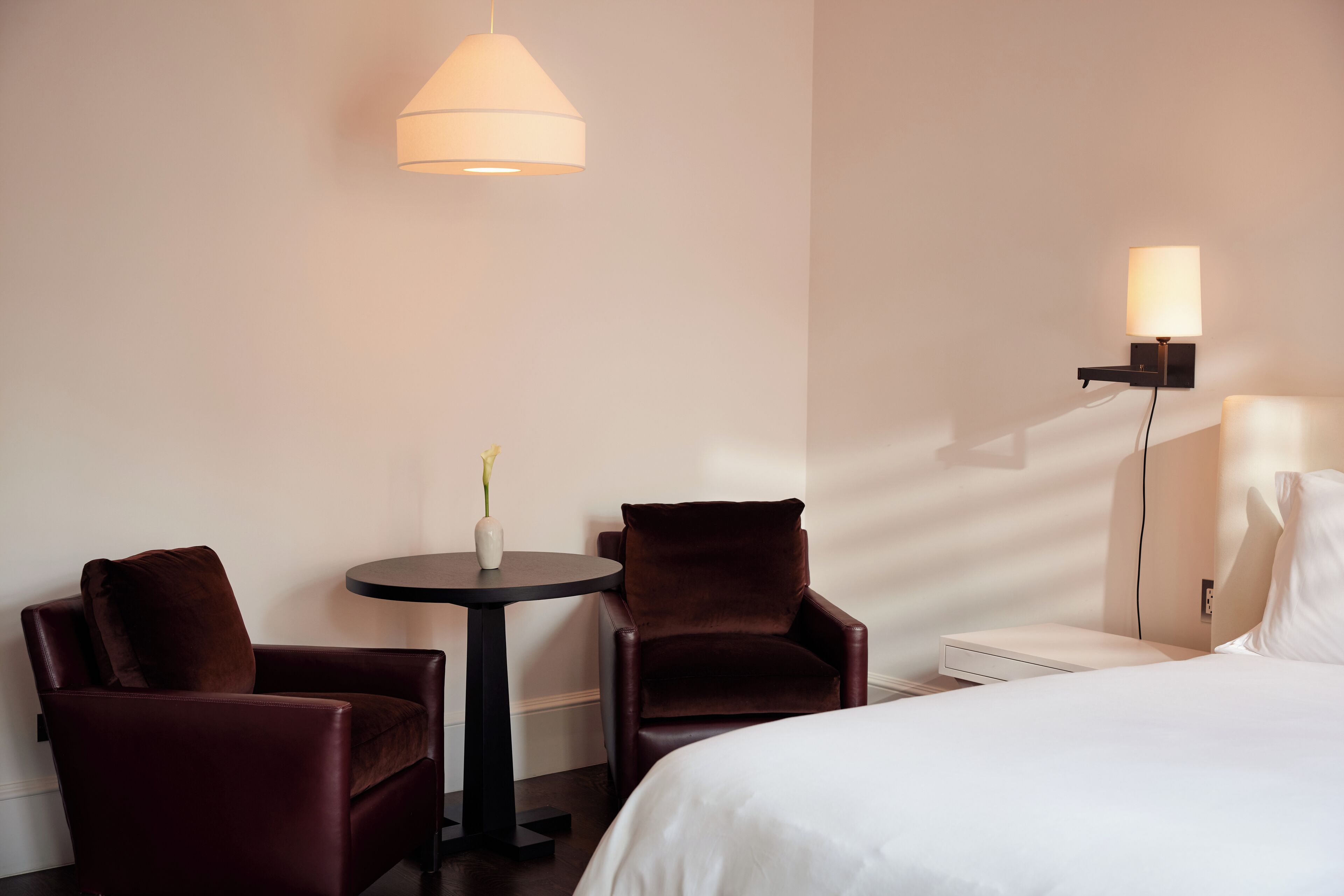 Una habitación de hotel con una cama, dos sillones, una mesita redonda con un florero y una lámpara de pared.