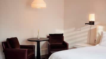 Una habitación de hotel con una cama, dos sillones, una mesita redonda con un florero y una lámpara de pared.