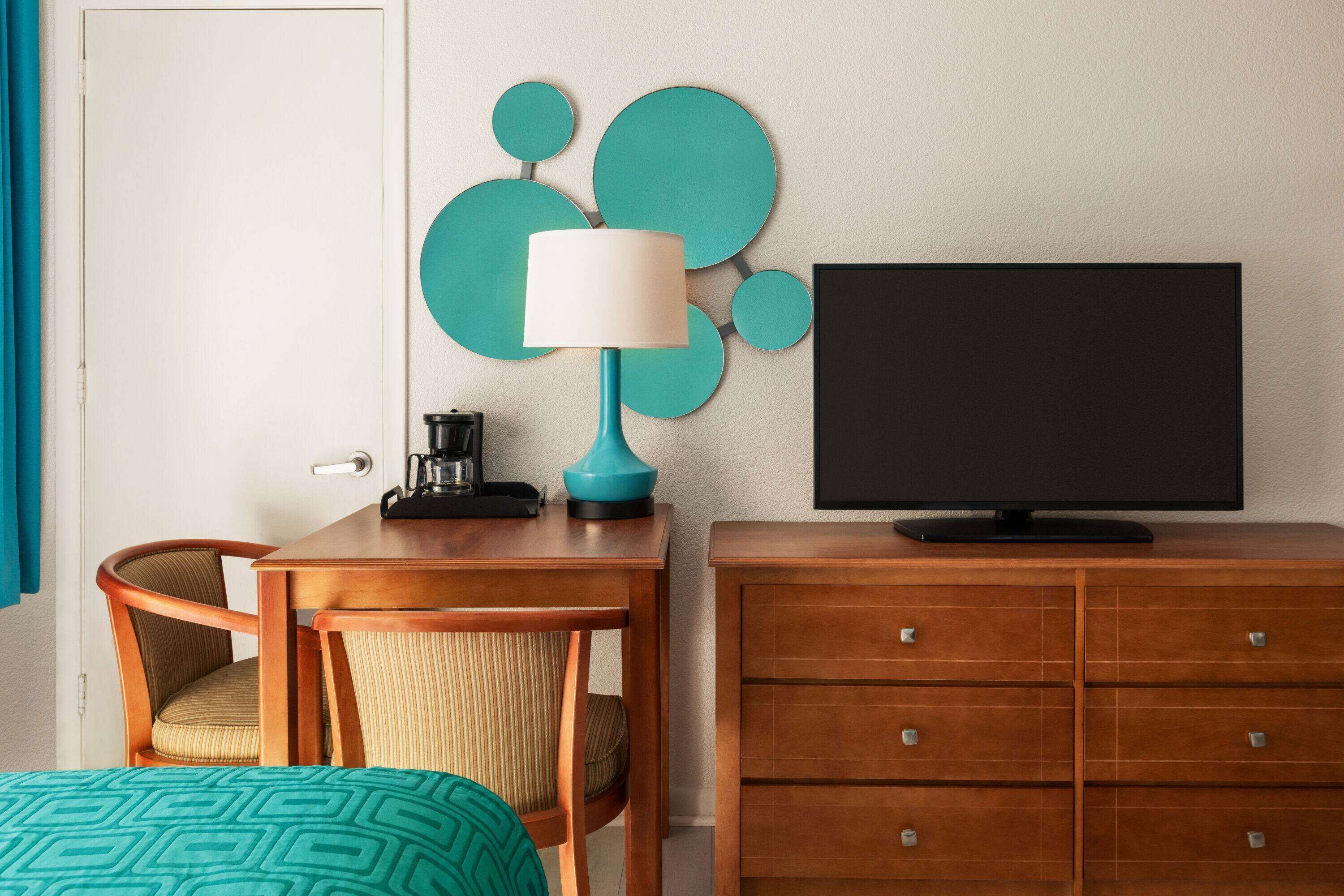 A hotel room with a wooden desk, a teal-patterned bed, a television on a wooden dresser, and a wall-mounted teal decorative piece.