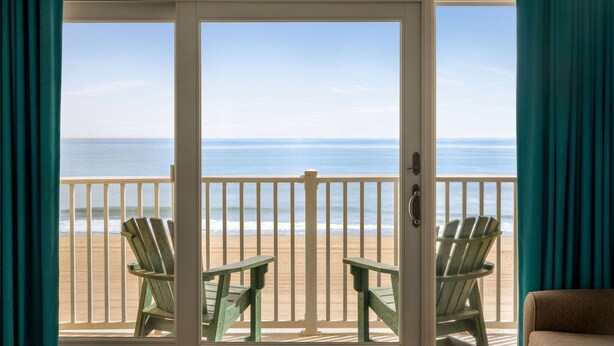 Una habitación con vistas a la playa a través de un ventanal, dos sillas Adirondack verdes y un sofá beige.
