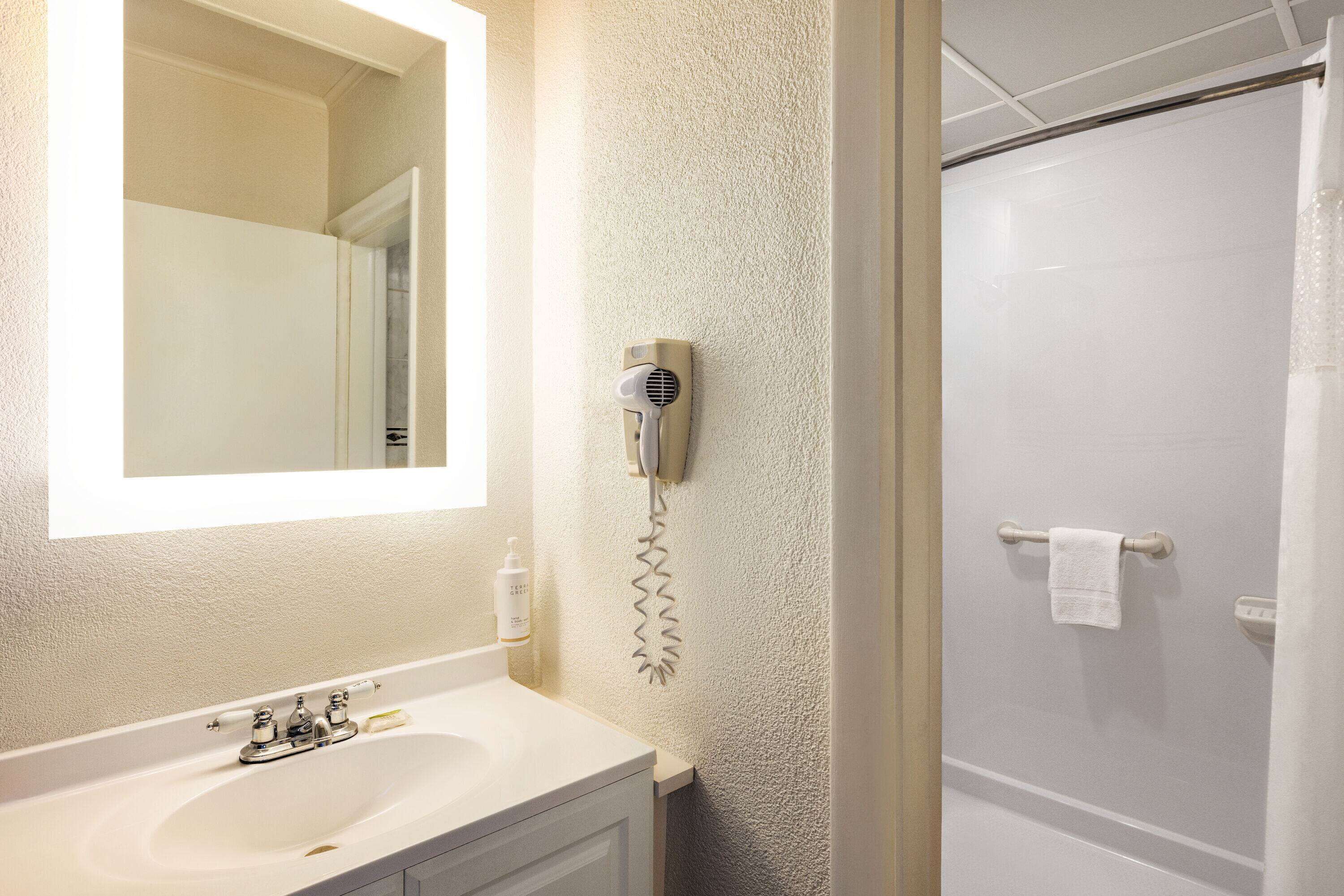 A bathroom with a white sink, a mirror, a hairdryer mounted on the wall, and a shower area.