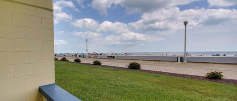 A view of a beach with a paved walkway, a lamp post, and a grassy area in the foreground.