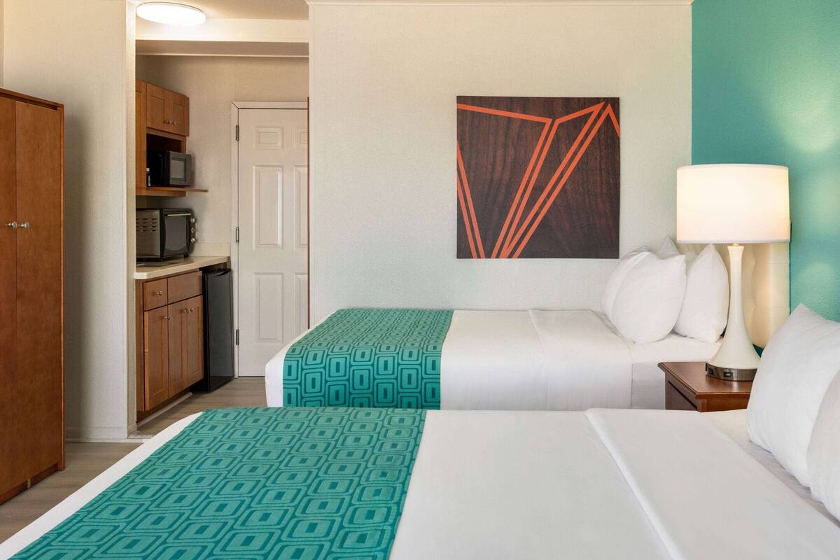 A hotel room with two beds, a kitchenette, a painting on the wall, and a bedside lamp.
