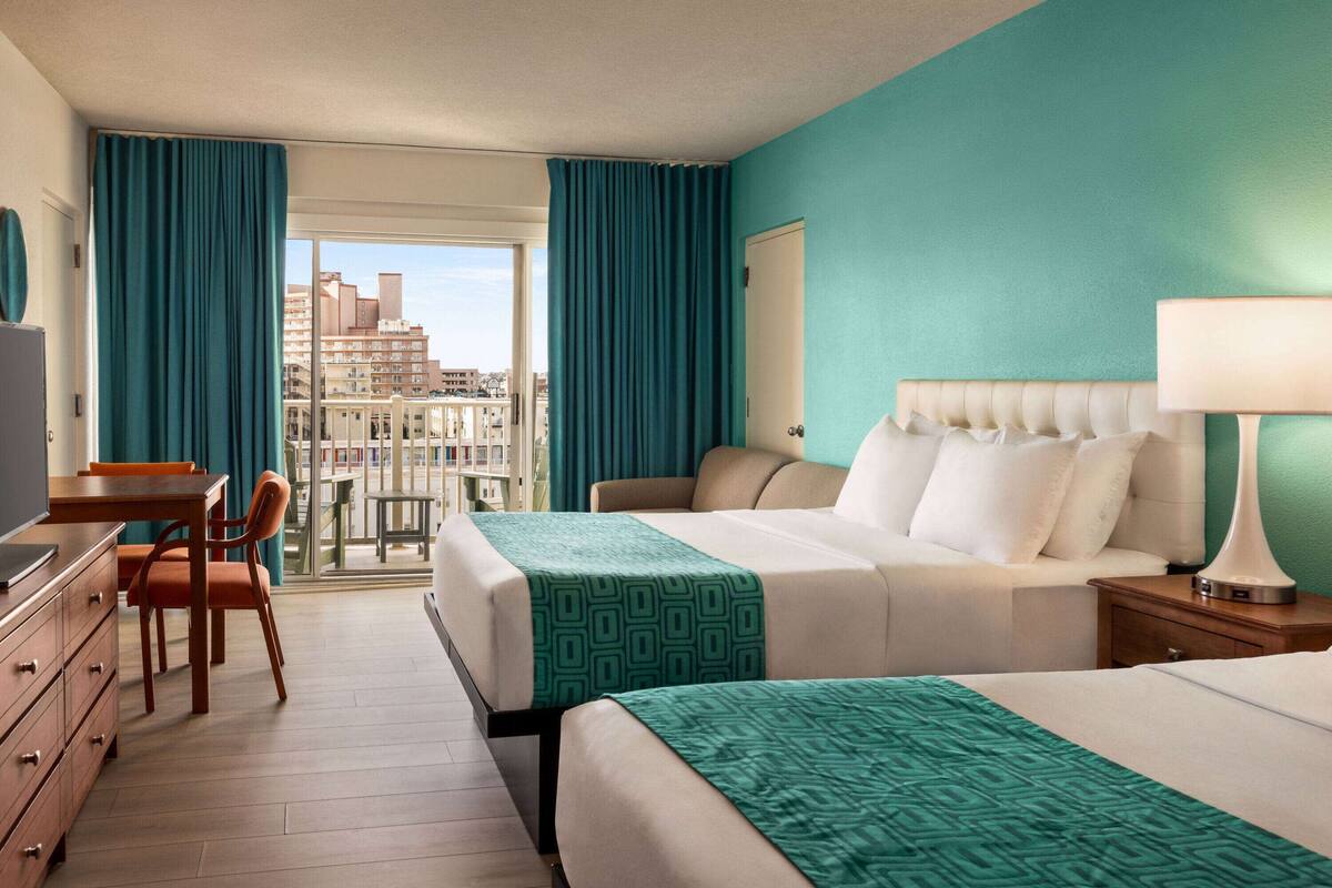 A hotel room with a bed, a desk, a chair, a dresser, and a view of the city through a large window.