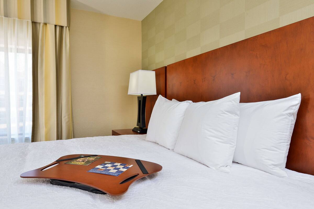 A hotel room with a neatly made bed, a wooden headboard, a nightstand with a lamp, and a window with curtains.