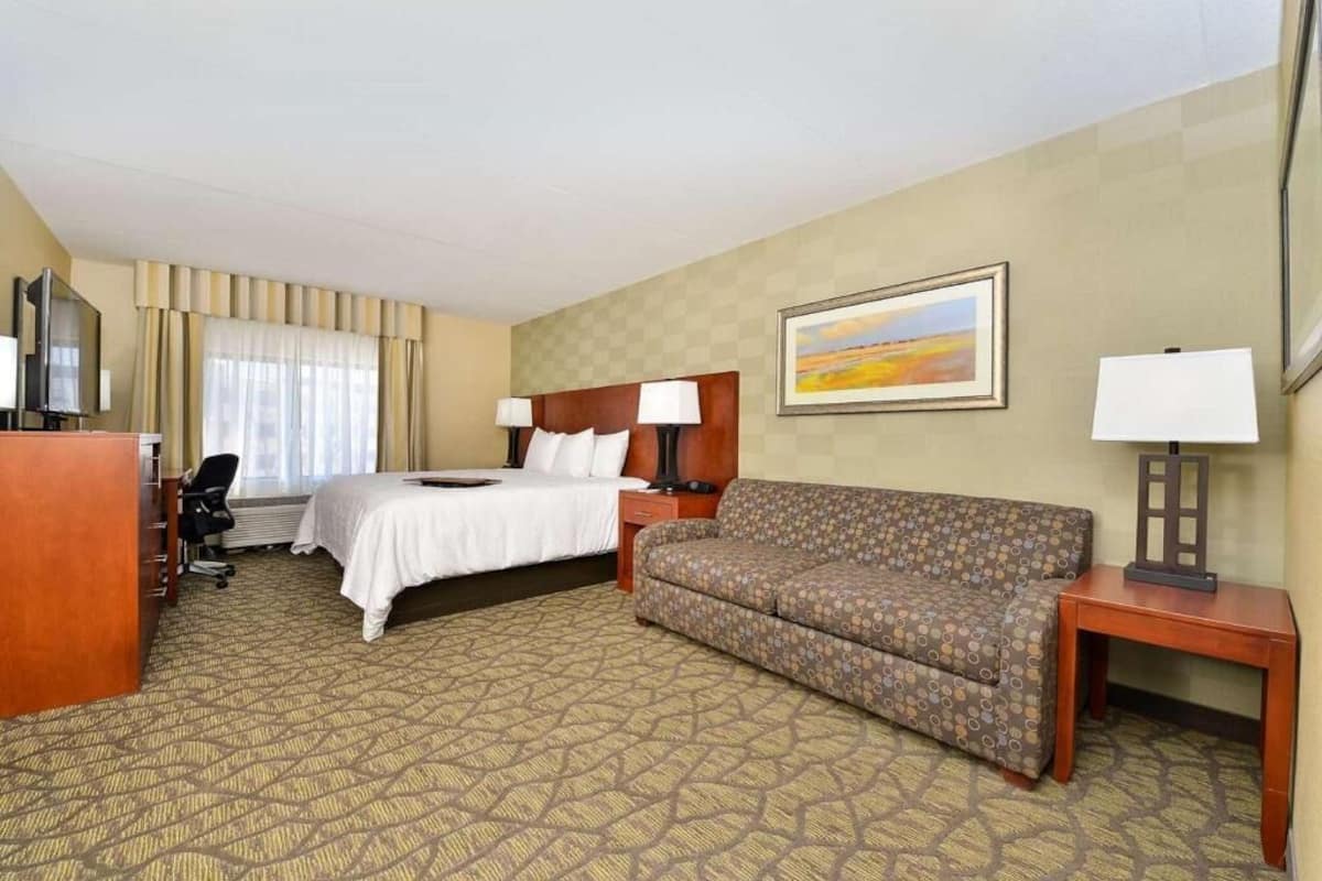 A hotel room with a bed, a sofa, a desk with a computer, a TV, and a painting on the wall.