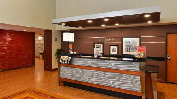 A reception desk with a wavy design, a welcome sign, framed pictures, and a red textured wall. - Hampton Inn Detroit/Auburn Hills South (Auburn Hills)