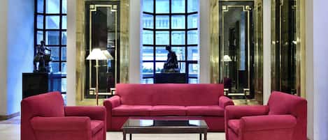 Un hall con un sofá rojo, una mesa de centro de vidrio y ventanas grandes con marcos decorativos.