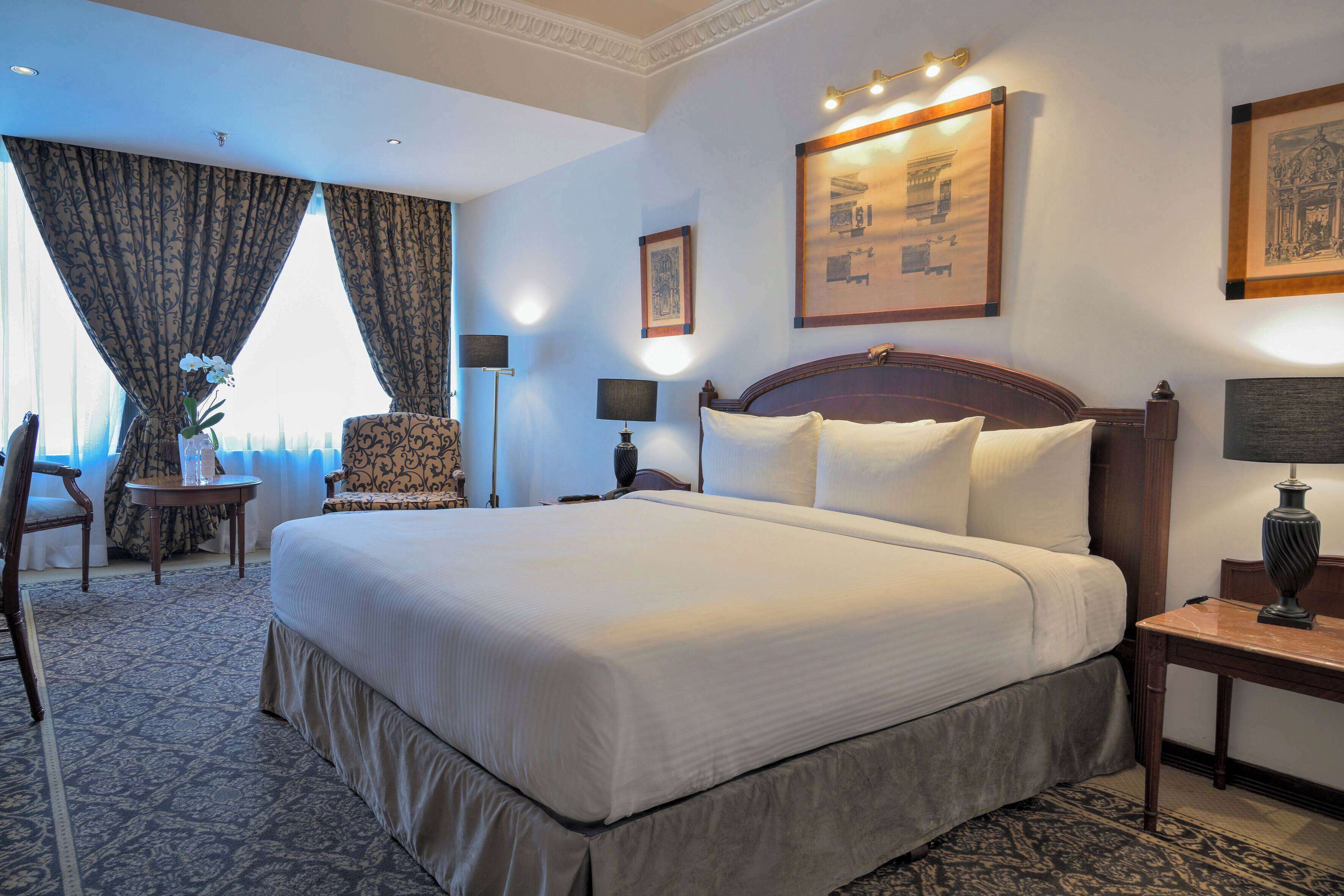A large bed with white linens, a wooden headboard, and a nightstand with a lamp. There is a chair with a patterned cushion and a small table with a vase of flowers. The room has a carpeted floor and a large window with curtains.