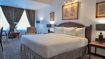 A large bed with white linens, a wooden headboard, and a nightstand with a lamp. There is a chair with a patterned cushion and a small table with a vase of flowers. The room has a carpeted floor and a large window with curtains.