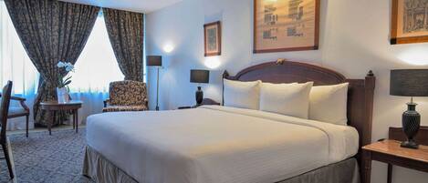 A large bed with white linens, a wooden headboard, and a nightstand with a lamp. There is a chair with a patterned cushion and a small table with a vase of flowers. The room has a carpeted floor and a large window with curtains.