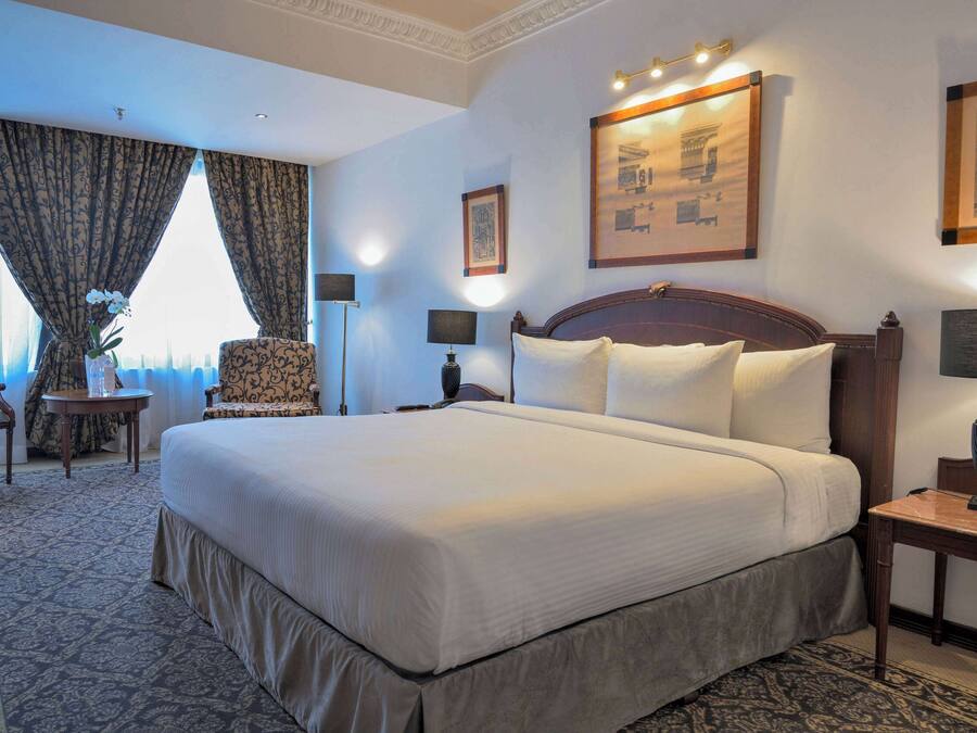 A large bed with white linens, a wooden headboard, and a nightstand with a lamp. There is a chair with a patterned cushion and a small table with a vase of flowers. The room has a carpeted floor and a large window with curtains.