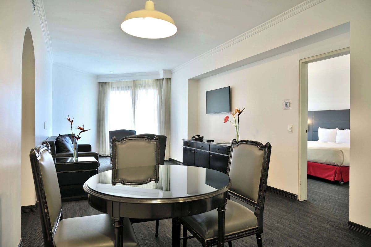 A hotel room with a round dining table, four chairs, a TV mounted on the wall, a bed with a red skirt, and a window with curtains.