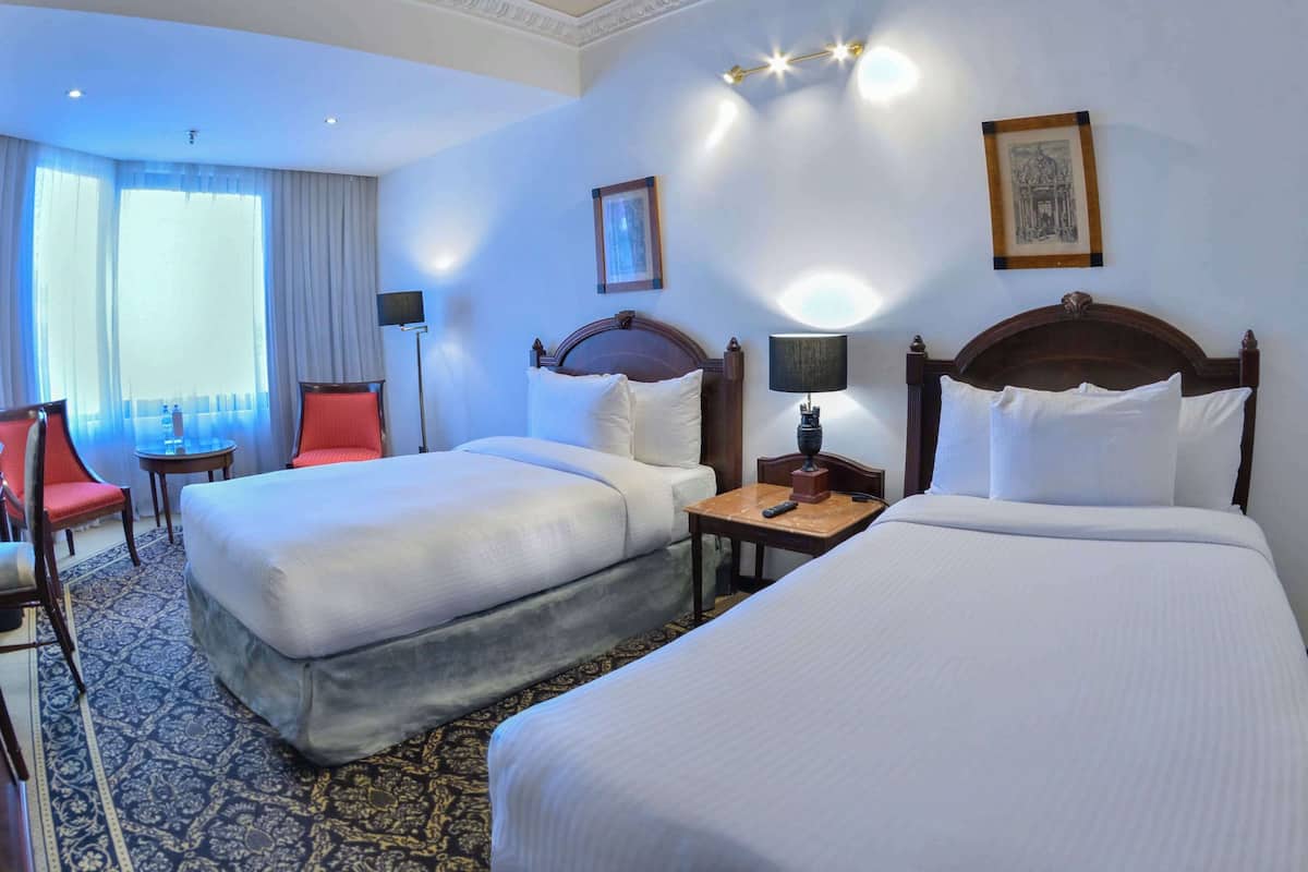 A hotel room with two beds, a red chair, a small table with a lamp, and a framed picture on the wall.