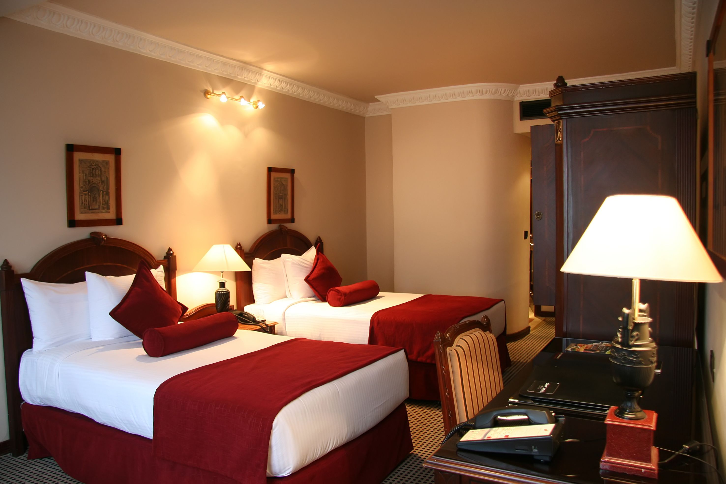 Ein Hotelzimmer mit zwei Betten, jeweils mit weißen Laken und roten Akzenten, einem Holzkopfstück, einem Schreibtisch mit Lampe und einem Spiegel.
