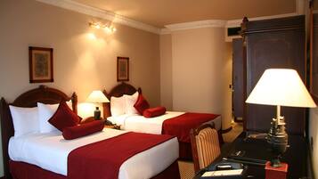 Ein Hotelzimmer mit zwei Betten, jeweils mit weißen Laken und roten Akzenten, einem Holzkopfstück, einem Schreibtisch mit Lampe und einem Spiegel.