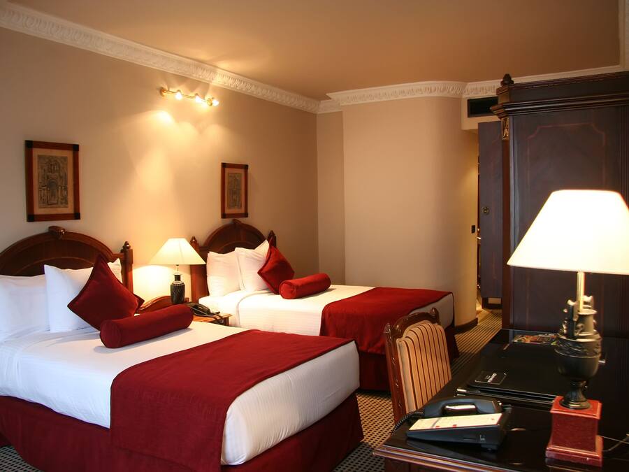 Ein Hotelzimmer mit zwei Betten, jeweils mit weißen Laken und roten Akzenten, einem Holzkopfstück, einem Schreibtisch mit Lampe und einem Spiegel.