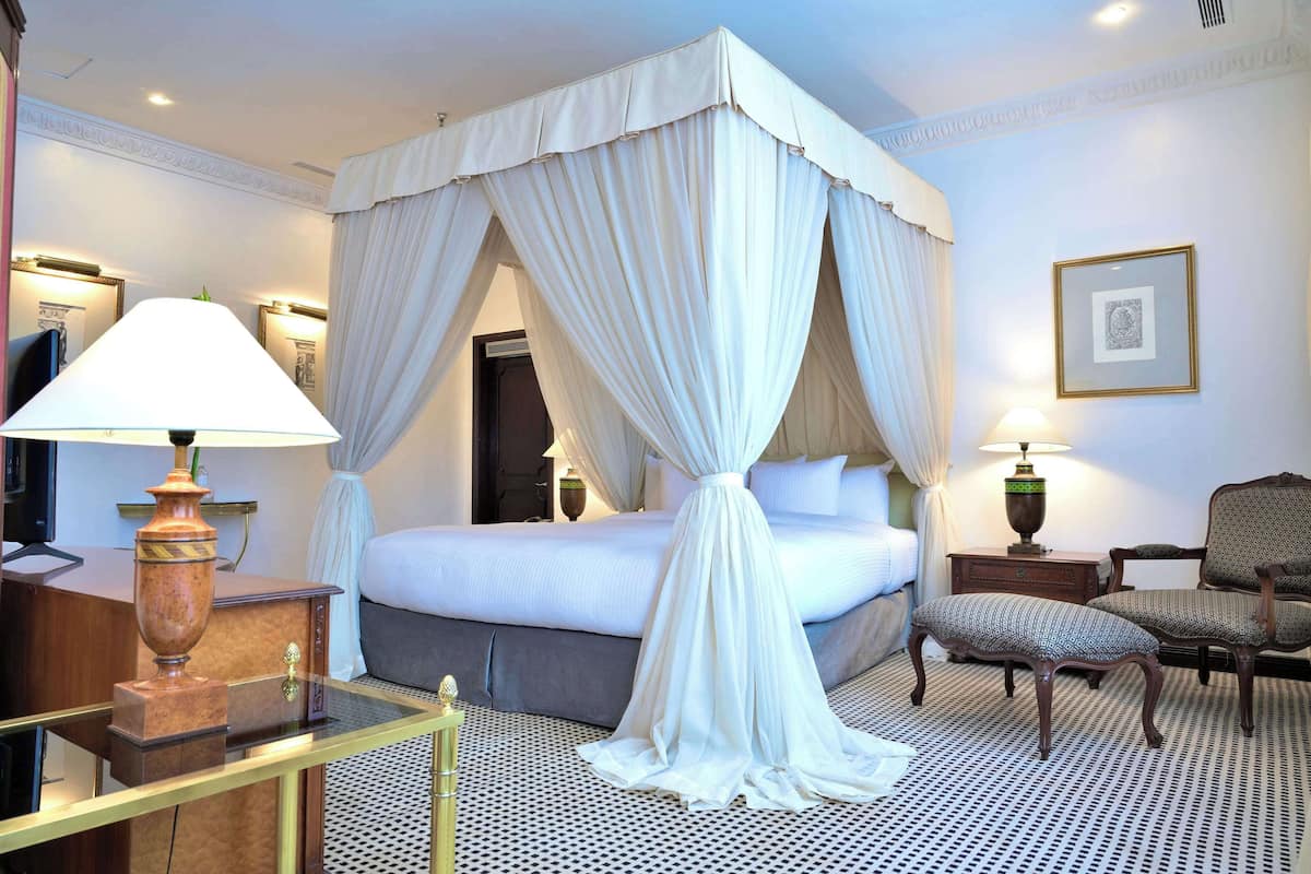 A hotel room with a four-poster bed, a desk with a lamp, a chair, and a patterned floor.