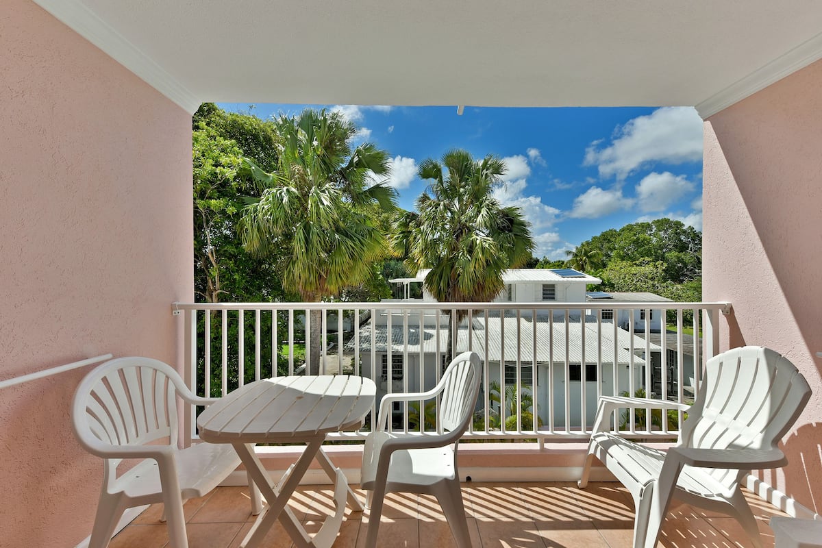 A balcony with white chairs and a table, overlooking a residential area with palm trees and a house.