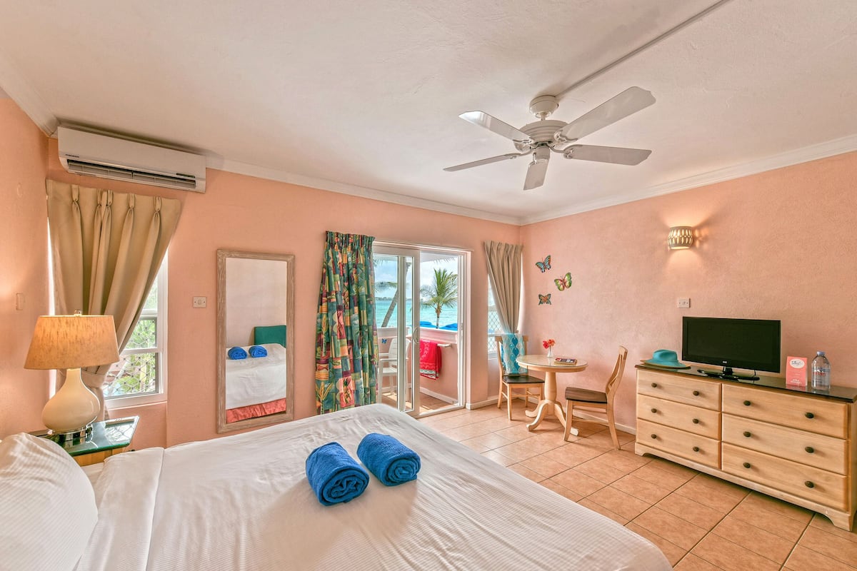 A hotel room with a bed, a ceiling fan, an air conditioner, a TV, a dresser, a chair, a table, and a balcony with a view.