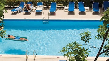 A swimming pool with a person on a floating board, surrounded by lounge chairs and a tiled deck.