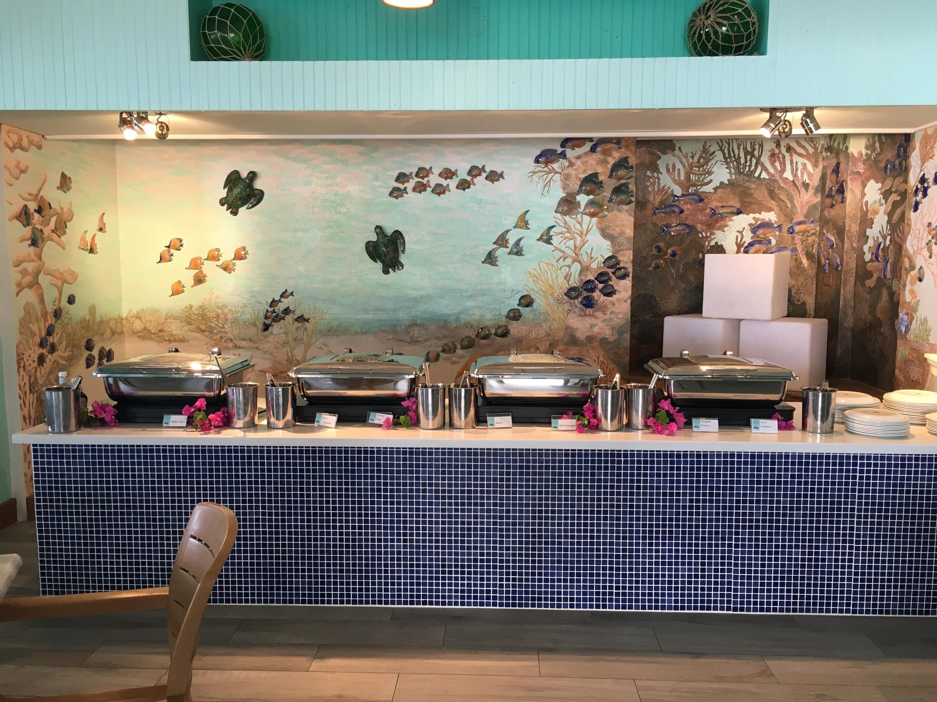 A buffet setup with multiple serving stations, a mural with aquatic and desert themes, and a tiled backsplash.