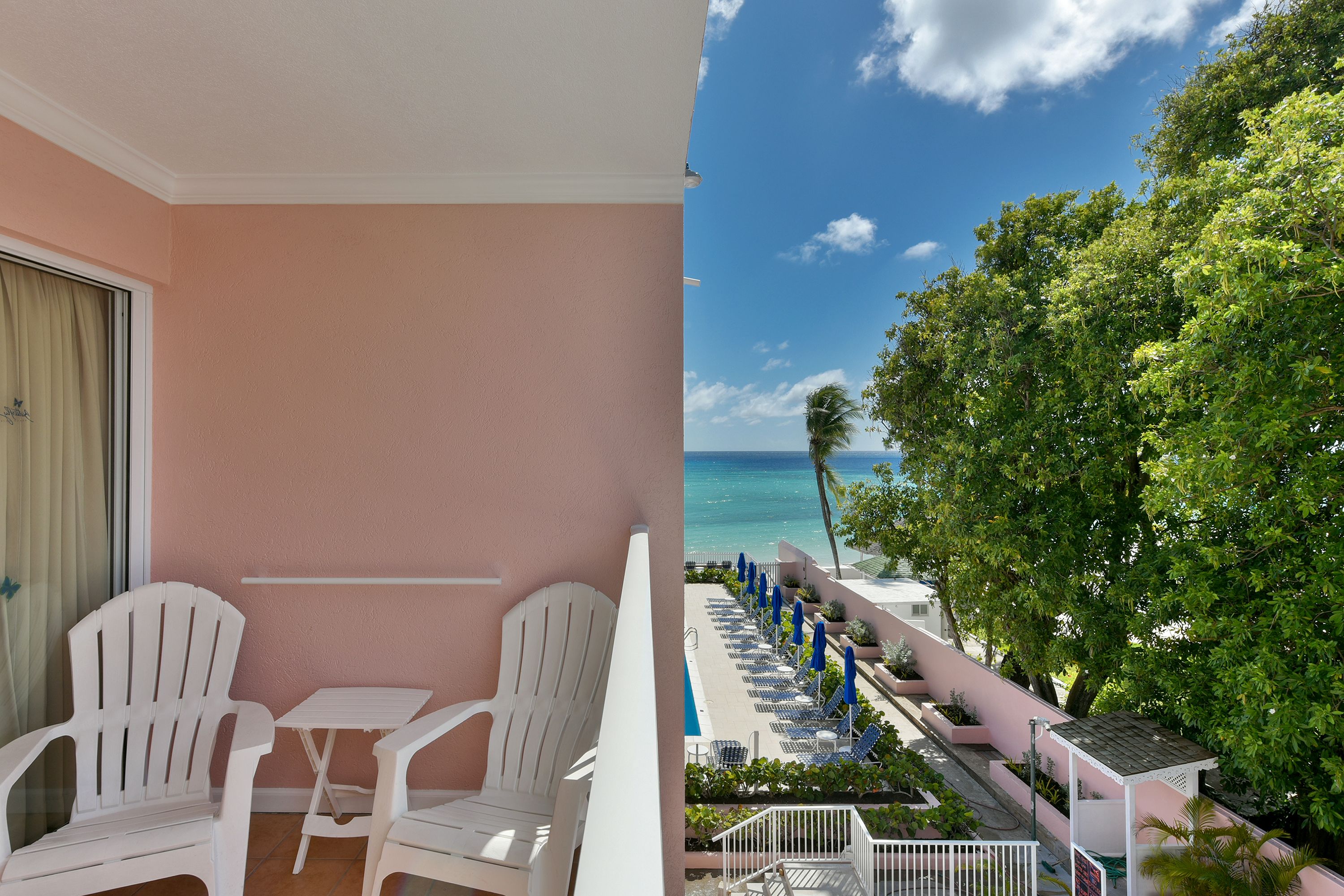 A balcony with white chairs and a table, overlooking a pool area with blue umbrellas and a view of the ocean.