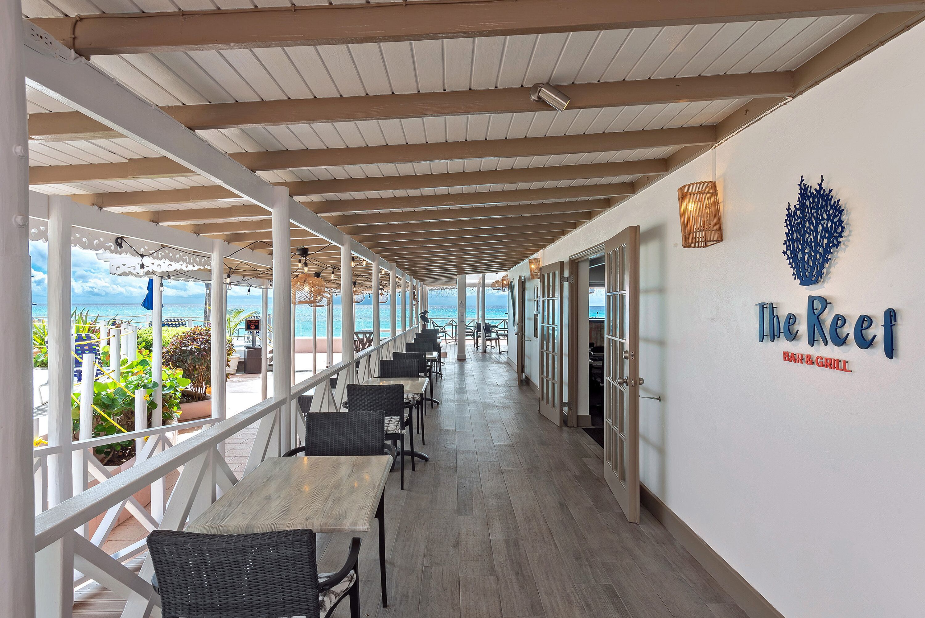 Et overdækket udendørs opholdsområde med borde og stole, udsigt til havet og et skilt med teksten "The Reef Bar & Grill".