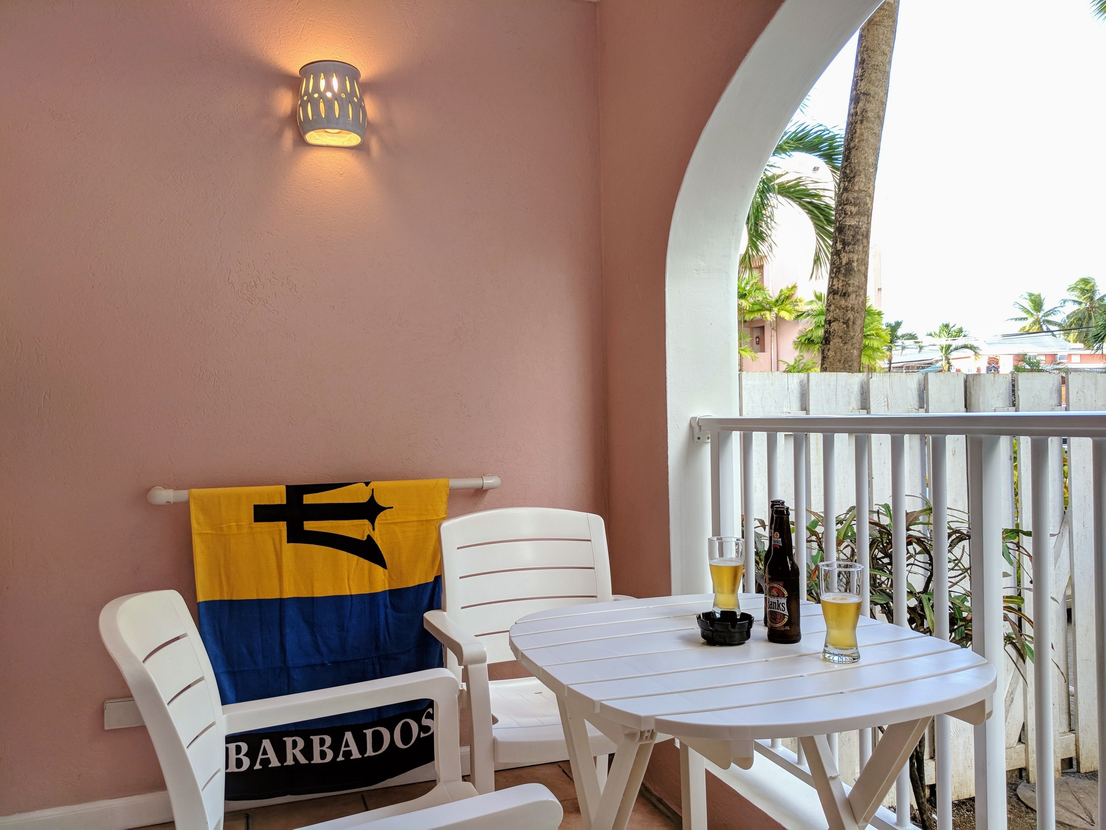 Un balcon avec une table blanche, deux chaises blanches, une serviette jaune et bleue, une bouteille de bière et un verre de bière.