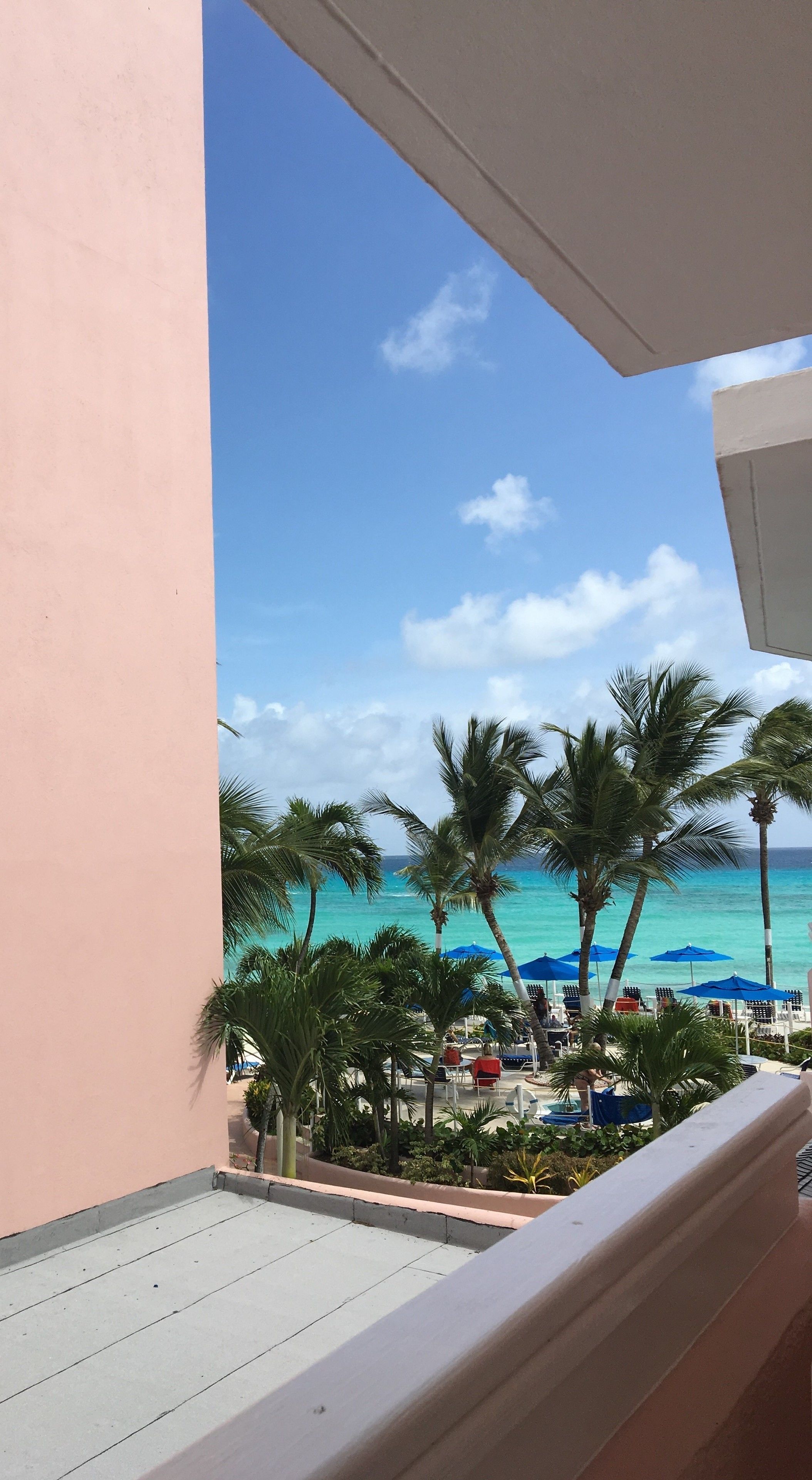 Un balcón con vista a una playa, palmeras y sombrillas azules.