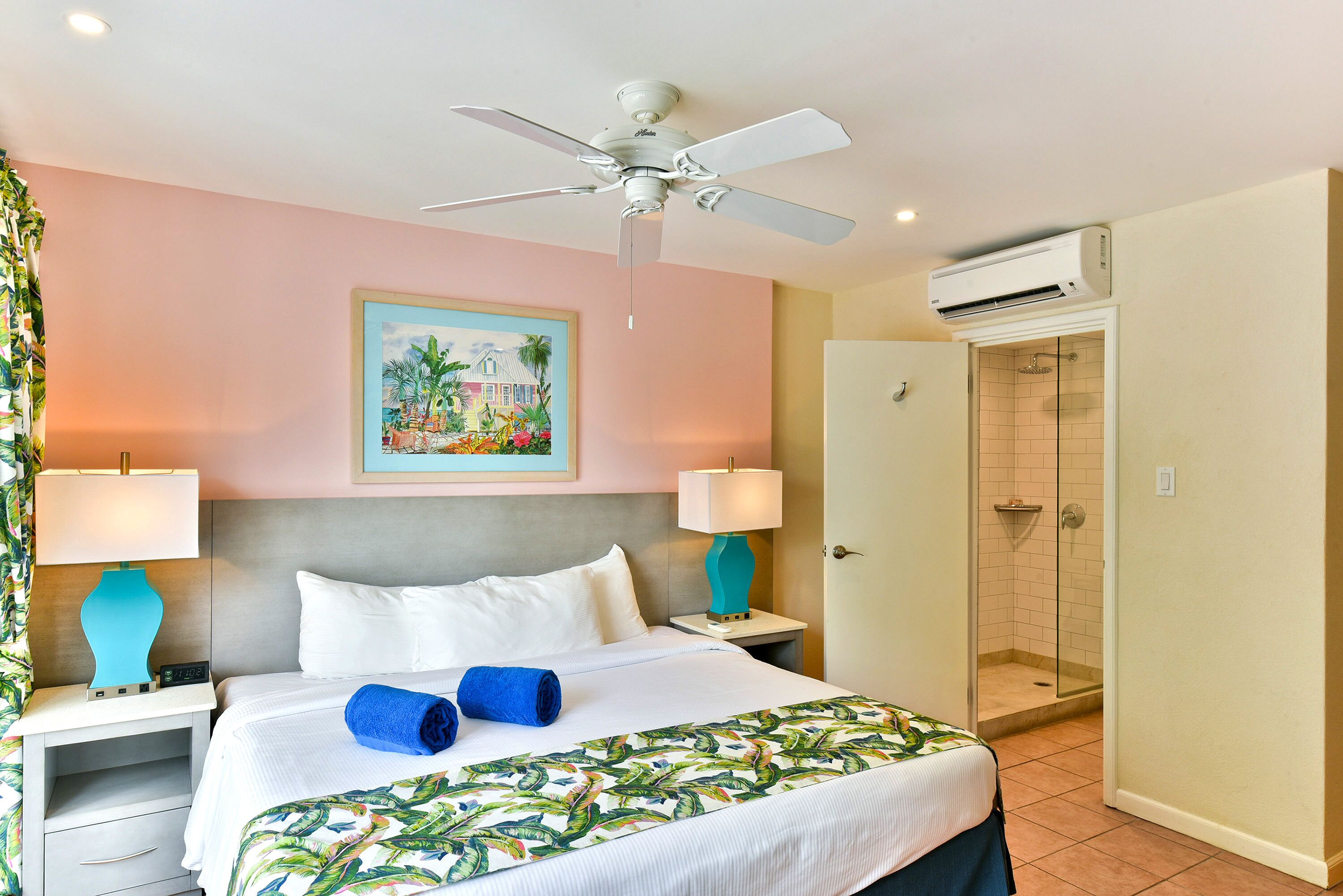 Habitación de hotel con cama, dos mesitas de noche, ventilador de techo, unidad de aire acondicionado, ducha y un cuadro en la pared.