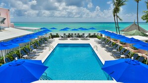 A pool area with blue umbrellas, lounge chairs, and a view of the ocean. - Butterfly Beach Hotel (Oistins)