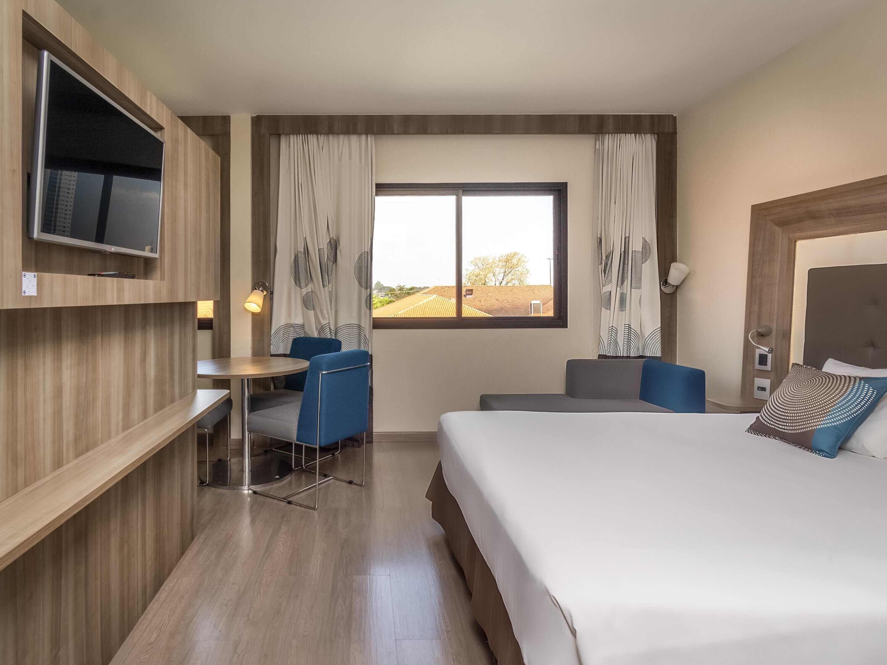 A hotel room with a bed, a desk, a chair, a TV, and a window with curtains.