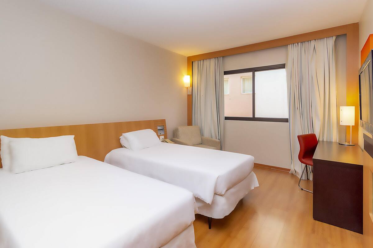 A hotel room with two beds, a desk with a chair, a lamp, and a window with curtains.