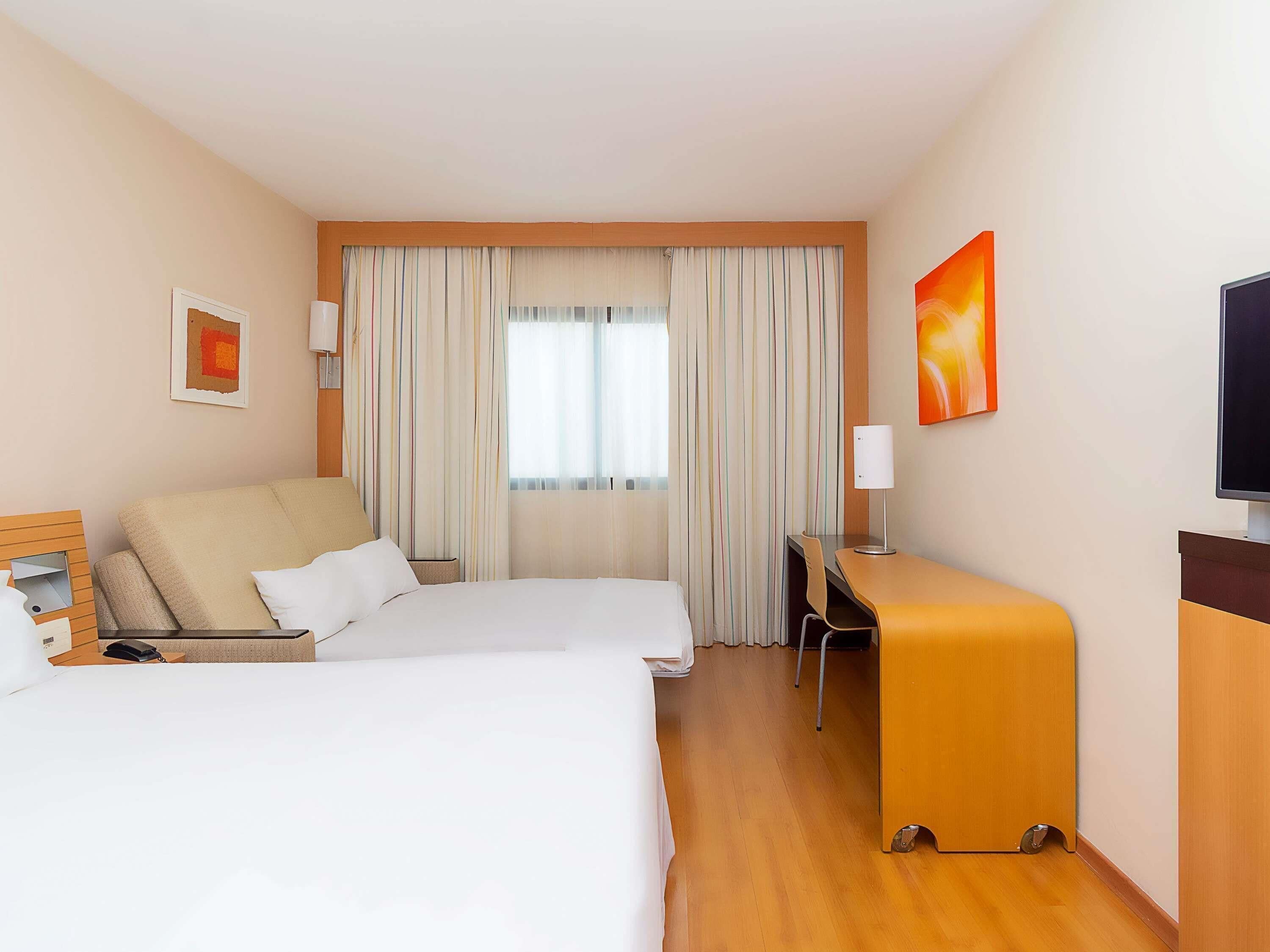A hotel room with two beds, a desk, a chair, a TV, and a window with curtains.