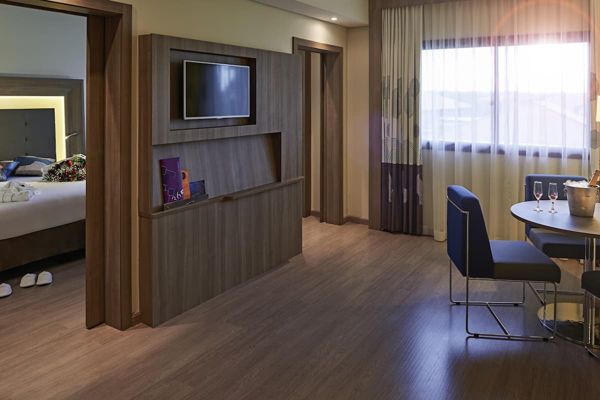 A hotel room with a bed, a TV mounted on the wall, a small table with two chairs, and a window with curtains.