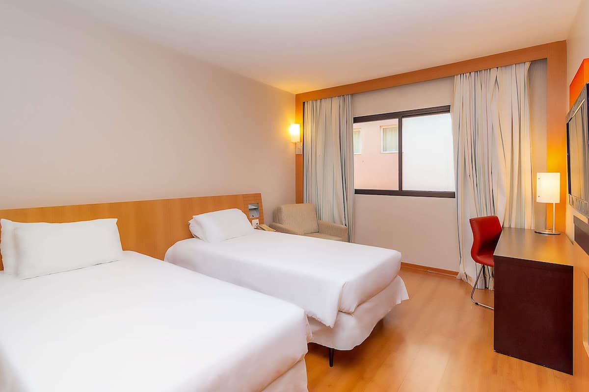 Ein Hotelzimmer mit zwei Betten, einem Schreibtisch, einem Stuhl, einem Fernseher und einem Fenster mit Vorhängen.